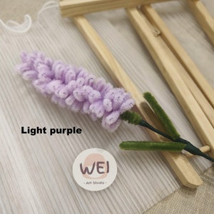 May include: Light purple artificial flower with green stem, displayed on a wooden easel with a white background. The flower is made of fuzzy material and resembles a lavender bloom. The background features a white fabric with a subtle striped pattern. A small white circle with the text "WEI - Art Studio -" is visible in the lower right corner.
