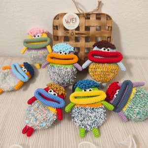 Può includere: Sette giocattoli di peluche all'uncinetto in vari colori e motivi. Ogni giocattolo ha una grande bocca colorata e un design unico. I giocattoli sono disposti su una superficie di legno con un cesto intrecciato sullo sfondo.