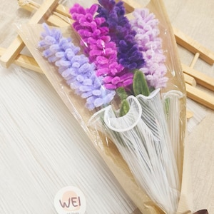 May include: A bouquet of purple, pink, and lavender pipe cleaner flowers wrapped in brown paper with a white ribbon and a brown ribbon with the text "love you" on it. The bouquet is labeled "WEI Art Studio."