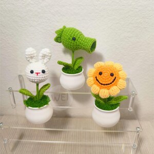 May include: Three crocheted plant figurines in white pots on a clear acrylic shelf. The figurines are a white bunny, a green plant with a mouth, and a yellow sunflower with a smiley face.