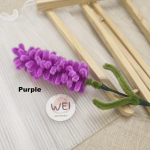 May include: Purple pipe cleaner flower with green stem and leaves, displayed on a wooden frame with white fabric behind it. The flower is made by Wei Art Studio.