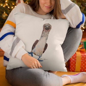 May include: A white throw pillow with a watercolor illustration of a blue-footed booby bird. The bird is facing forward with its head tilted slightly to the side. The bird's feet are blue and its beak is orange. The pillow is being held by a person.