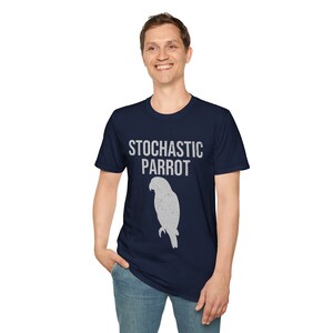 May include: A navy blue short-sleeved t-shirt featuring the white text STOCHASTIC PARROT printed in a distressed font above a white silhouette graphic of a parrot. The design is centered on the chest of the garment, which has a crew neckline.