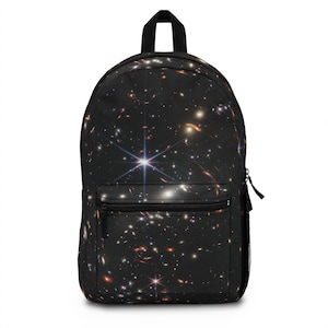 Puede incluir: Mochila negra con estampado de galaxia que presenta una estrella blanca brillante y otras estrellas en tonos naranja, amarillo y azul.