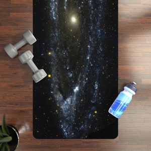 May include: A black yoga mat with a galaxy print featuring a swirling nebula and stars. The mat is on a wooden floor with two gray dumbbells and a blue water bottle.