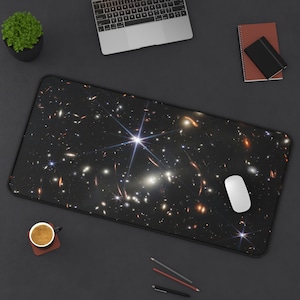 May include: A black mouse pad with a galaxy print featuring a bright star in the center. The mouse pad is on a dark gray surface with a laptop, a cup of coffee, pencils, and a notebook.