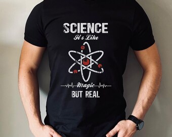 Può includere: Una t-shirt nera a maniche corte con una grafica bianca di un atomo con accenti rossi. Il testo "SCIENCE It's Like magic BUT REAL" è stampato in un carattere bianco effetto usurato, con una linea di battito cardiaco sotto l'atomo.
