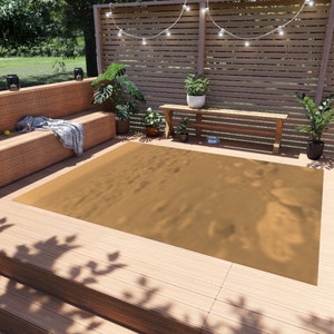 May include: A tan outdoor rug with a subtle textured pattern. The rug is laid out on a wooden deck with a bench and plants in the background.