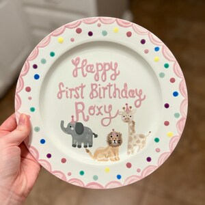 Puede incluir: Un plato de cer&aacute;mica blanco con un borde festoneado rosa y lunares de colores. El plato presenta el texto "Happy First Birthday Roxy" en rosa, junto con ilustraciones de un elefante, un le&oacute;n y una jirafa, todos con sombreros de fiesta.