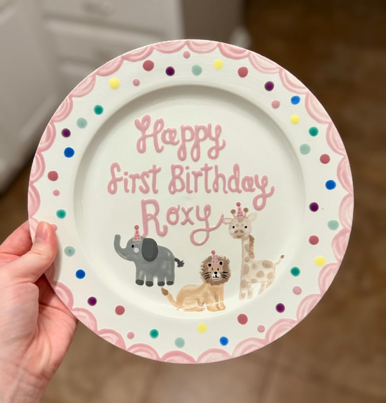 Puede incluir: Un plato de cer&aacute;mica blanco con un borde festoneado rosa y lunares de colores. El plato presenta el texto "Happy First Birthday Roxy" en rosa, junto con ilustraciones de un elefante, un le&oacute;n y una jirafa, todos con sombreros de fiesta.