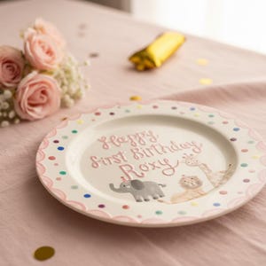 Puede incluir: Un plato de cer&aacute;mica blanco con borde festoneado rosa y lunares de colores. El plato presenta el texto "Happy First Birthday Roxy" e ilustraciones de un elefante, un le&oacute;n y una jirafa. Un ramo de rosas rosas y confeti dorado est&aacute;n cerca.