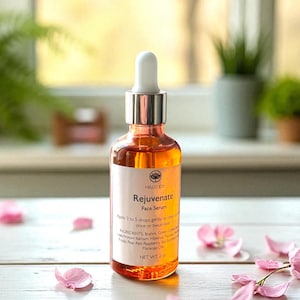 May include: A clear glass bottle of Hello Joy Rejuvenate Face Serum with a white dropper. The orange serum is labelled with the product name and ingredients. Pink flower petals are scattered around the bottle on a white wooden surface.