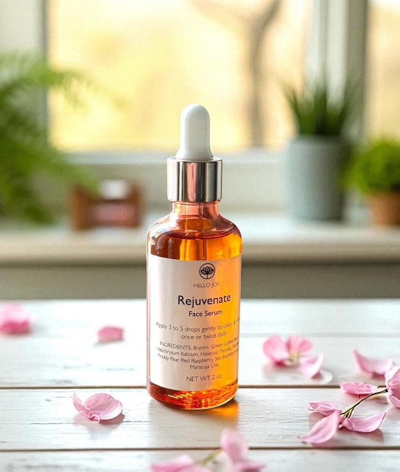 May include: A clear glass bottle of Hello Joy Rejuvenate Face Serum with a white dropper. The orange serum is labeled with the product name and ingredients. Pink flower petals are scattered around the bottle on a white wooden surface.