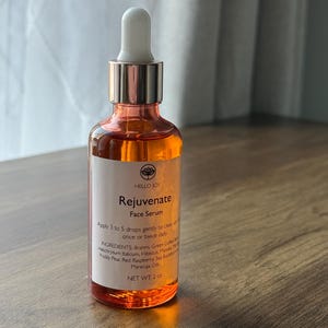 May include: A clear glass bottle of Hello Joy Rejuvenate Face Serum. The bottle has a white dropper and a rose gold cap. The label includes the product name, instructions, ingredients, and net weight of 2 oz.