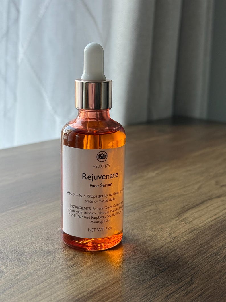 May include: A clear glass bottle of Hello Joy Rejuvenate Face Serum. The bottle has a white dropper and a rose gold cap. The label includes the product name, instructions, ingredients, and net weight of 2 oz.