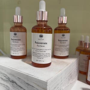 May include: Three amber glass bottles of Hello Joy Rejuvenate Face Serum with white labels and rose gold droppers. The labels list ingredients and usage instructions. Each bottle contains 2 oz of product.