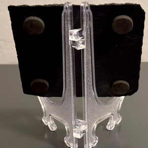 May include: A black slate board with four brown circular magnets on the back, supported by a clear plastic easel. The easel has a decorative design and is made of clear plastic. The slate board is rectangular with a rough, textured surface.