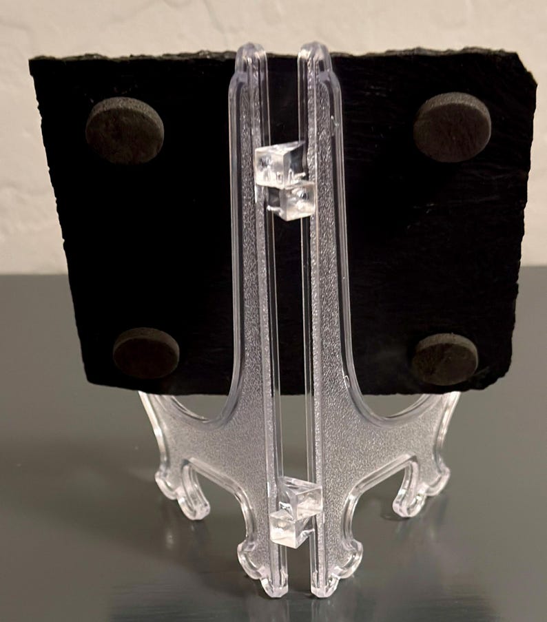 May include: A black slate board with four brown circular magnets on the back, supported by a clear plastic easel. The easel has a decorative design and is made of clear plastic. The slate board is rectangular with a rough, textured surface.