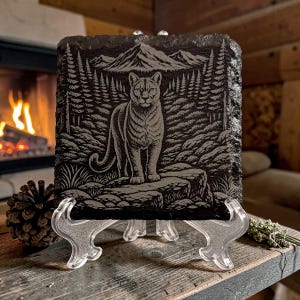 May include: A rectangular, dark grey slate tile with an engraved cougar standing on a rock, set against a forest and mountain backdrop. The tile is displayed on a clear plastic stand, showcasing the detailed wildlife art. The theme is nature and wildlife.