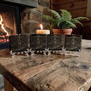 May include: Four square, dark coasters featuring a tiger design, presented on clear plastic stands. They are arranged on a rustic wooden table, with a lit candle, a potted fern, and a fireplace in the background.