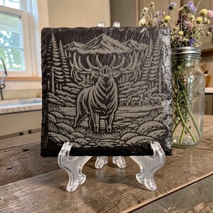 May include: A square, dark grey slate tile featuring an engraved image of a stag in a forest scene. The stag has large antlers and is surrounded by trees, mountains, and other deer. The tile is displayed on a clear plastic stand.
