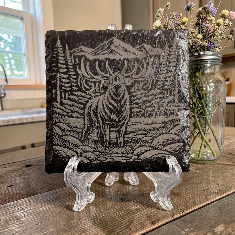 May include: A square, dark grey slate tile featuring an engraved image of a stag in a forest scene. The stag has large antlers and is surrounded by trees, mountains, and other deer. The tile is displayed on a clear plastic stand.