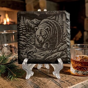 May include: A square, dark grey slate coaster featuring an engraved image of a bear in a forest scene, holding a fish. The coaster is displayed on a clear plastic stand, with a glass of amber liquid and a fireplace in the background.