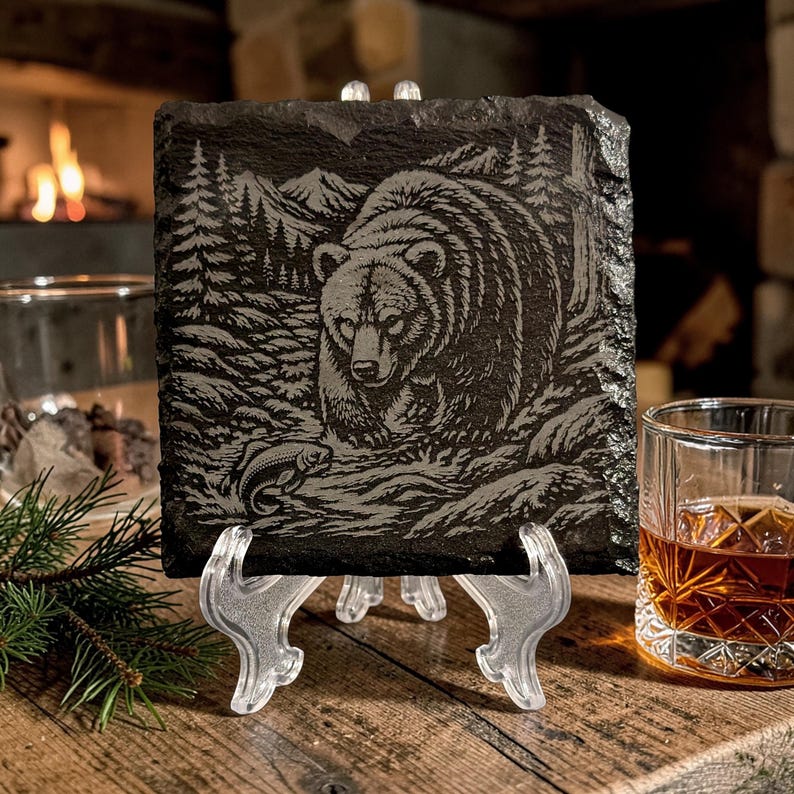 May include: A square, dark grey slate coaster featuring an engraved image of a bear in a forest scene, holding a fish. The coaster is displayed on a clear plastic stand, with a glass of amber liquid and a fireplace in the background.