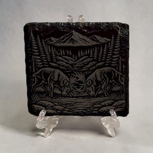 May include: A black slate coaster featuring an etched forest scene. Two deer are fighting, with a mountain range and trees in the background. The coaster is supported by a clear plastic stand.