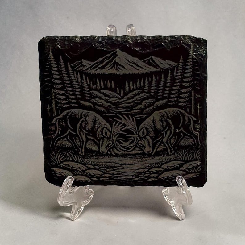 May include: A black slate coaster featuring an etched forest scene. Two deer are fighting, with a mountain range and trees in the background. The coaster is supported by a clear plastic stand.
