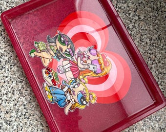 May include: A rectangular, burgundy-colored tray with a glittery finish. The tray features a graphic of three cartoon characters in the center, set against a red and pink target design. The characters are in various poses and outfits.