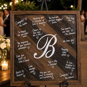 May include: A wooden wedding sign with a dark brown frame and a large white letter "B" in the center. The sign is decorated with handwritten messages and hearts, perfect for a wedding or engagement celebration.