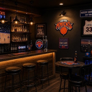 May include: A New York Knicks-themed bar area with a wooden bar, stools, and a television screen displaying a basketball game. The walls are decorated with Knicks memorabilia, including a jersey, logo, and sign that says "Knicks Fan Cave".