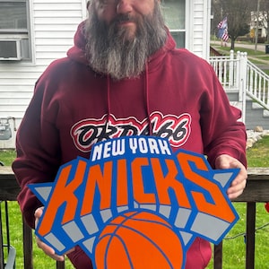 May include: A person holding a large, wooden New York Knicks logo. The logo features the team name in orange and blue, with a basketball in the center. The person is wearing a maroon hoodie with the team's name on it.