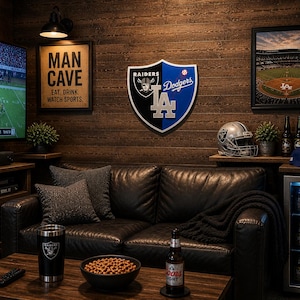 Puede incluir: Un interior de cueva de hombre con un sofá de cuero negro, una mesa de centro de madera y un televisor que muestra un partido de fútbol americano. La pared presenta un emblema de Raiders/Dodgers, un letrero de "Man Cave" y una impresión de estadio de béisbol.