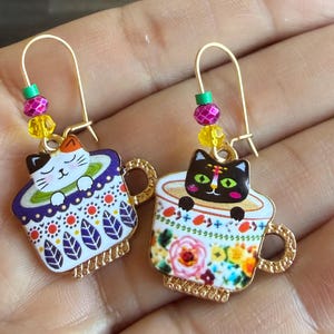 May include: A pair of gold-tone cat earrings. Each earring features a colorful teacup design with a cat peeking over the rim. The teacups are decorated with floral and geometric patterns. The earrings have beaded accents.