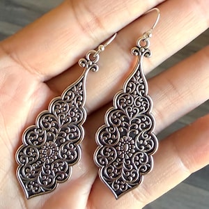 May include: A pair of silver-colored dangle earrings. Each earring features an ornate, symmetrical design with intricate scrollwork and floral patterns. The earrings have a teardrop shape and are suspended from a simple hook.