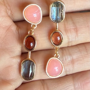 May include: A pair of gold-tone drop earrings featuring three vertically aligned stones each. The left earring has a peach, a dark red, and a dark gray stone. The right earring has a dark gray, a dark red, and a peach stone in a vertical row.