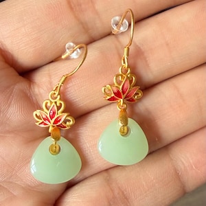 May include: A pair of gold-colored earrings with a red and gold lotus flower design and a light green, triangular jade stone. The earrings have a hook closure.