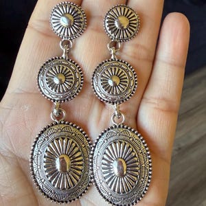 May include: Silver-toned, three-tiered dangle earrings. The earrings feature a circular top, a circular middle, and an oval bottom. Each tier has a sunburst design. The earrings are connected by small, curved links. The earrings are displayed against a neutral background.