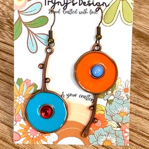 May include: A pair of handmade earrings with a floral design. One earring features a bright orange circle with a light blue center, while the other has a turquoise circle with a red center. The earrings are attached to a copper-colored stem.