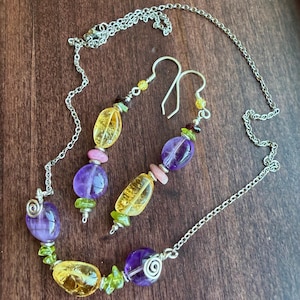 May include: A jewelry set consisting of a necklace and two drop earrings. The pieces feature polished yellow citrine, purple amethyst, and small green peridot beads strung on silver-toned wire and chain with spiral metal accents.