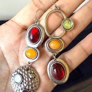 May include: Two silver-toned dangle earrings featuring a multi-layered design. One earring has a red cabochon, an orange cabochon, and a textured circular disc with two small inset stones. The other has a green cabochon, an orange cabochon, and a red cabochon.