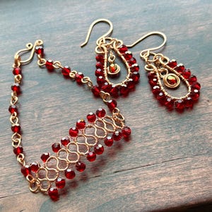 May include: A gold-toned wire bracelet and a pair of matching drop earrings, each featuring small, faceted, deep red crystal beads. The bracelet has a woven, link-style design, while the earrings consist of teardrop-shaped wire frames.