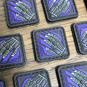 May include: A set of eight purple and black embroidered patches featuring a skeletal hand with claws. The patches are square and have a black border.