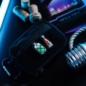 Könnte beinhalten: Eine schwarze Reißverschlusstasche mit zwei gestickten Aufnähern. Ein Aufnäher ist grün und zeigt eine Flasche mit grüner Flüssigkeit. Der andere Aufnäher ist orange und zeigt eine Flasche mit roter Flüssigkeit.