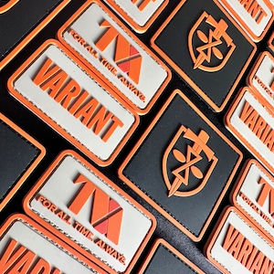 Puede incluir: Primer plano de varios parches rectangulares con la palabra "VARIANT" en naranja y el texto "FOR ALL TIME. ALWAYS.". Los parches tienen un fondo negro o blanco con diseños geométricos naranjas y rojos.