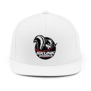 May include: A white baseball cap featuring a circular logo on the front. The logo depicts a black and white skunk illustration with a small jet silhouette, accompanied by the text SKUNK WORKS in bold white letters on a black background.
