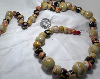 Puede incluir: Un collar con cuentas redondas en crema, negro, rojo y amarillo, de varios tamaños y con un acabado brillante. Se incluye una moneda como referencia de tamaño. El collar se muestra sobre una superficie blanca.