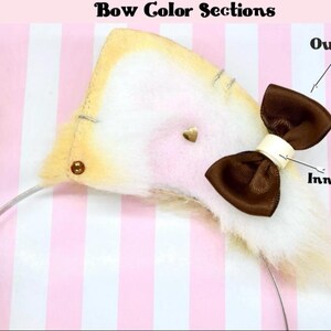 Puede incluir: Primer plano de una diadema de orejas de gato con un lazo marr&oacute;n. La oreja es beige con un interior blanco y un peque&ntilde;o coraz&oacute;n dorado. El texto "Bow Color Sections" est&aacute; en la parte superior. El fondo es de rayas rosas y blancas.